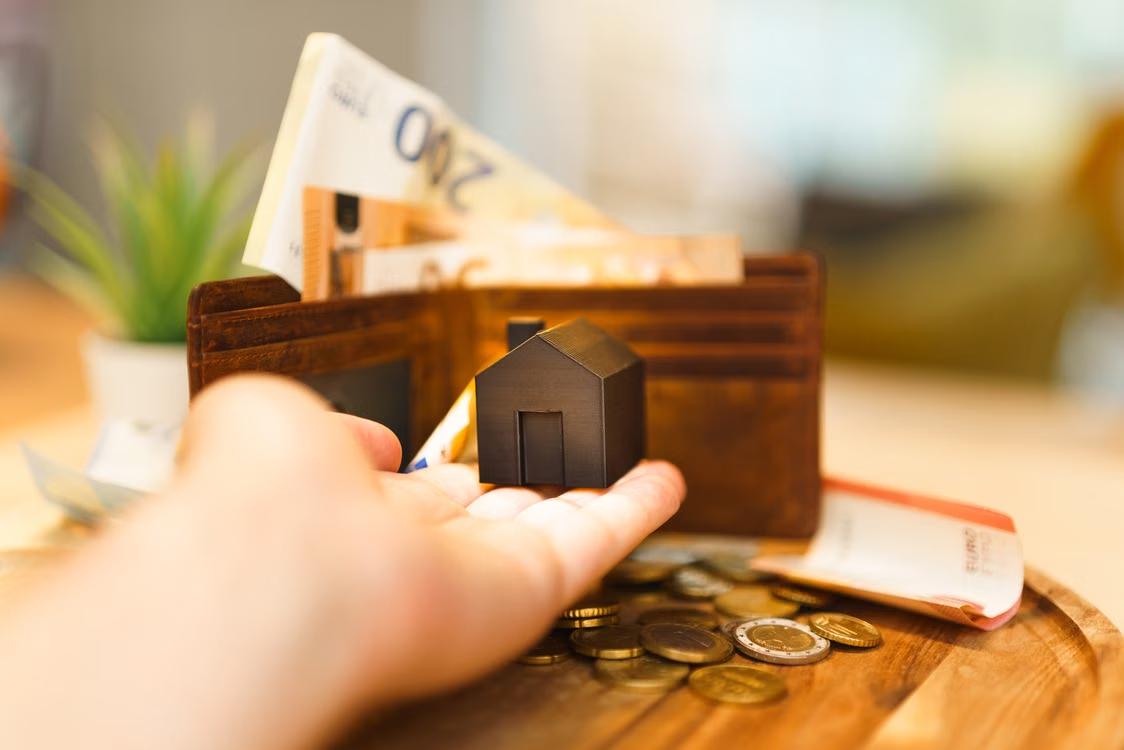 An image of a person holding a miniature house on a table with coins and money