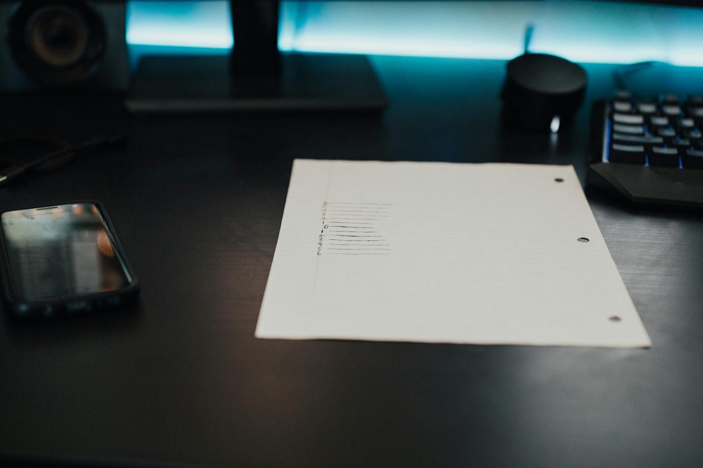Sheet of paper on a dark desk beside a smartphone and keyboard